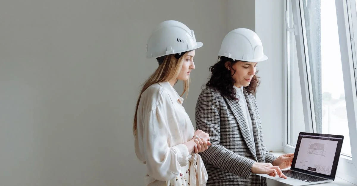 Deux professionnelles en casques de sécurité examinent des plans de construction sur un ordinateur portable près d'une fenêtre.