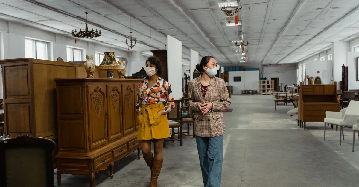 Deux femmes portant des masques explorent un grand magasin rempli de meubles vintage et de lustres.
